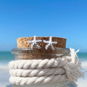 Dainty Pearl Starfish Stud Earrings displayed on a cork bottle against a blurred beach background.