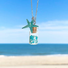 Load image into Gallery viewer, Glowing Sand Jar Necklace hanging close for a shot with a blurred beach background.