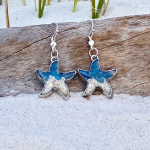 Sand Starfish Earrings in Blue Glass are displayed by being placed on top of a driftwood on the sand.