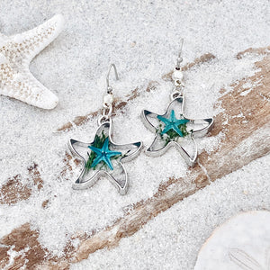 Deep In The Ocean Starfish Earrings are displayed by being placed on top of a sand covered driftwood.