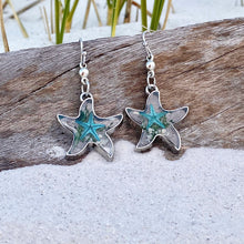 Load image into Gallery viewer, Deep In The Ocean Starfish Earrings are displayed by being placed on top of a driftwood on the sand.