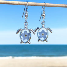 Load image into Gallery viewer, Forget Me Not Sea Turtle Earrings hanging on a stick against a blurred beach background, perfect for ocean-inspired jewelry.