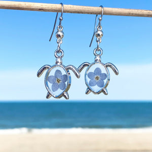 Forget Me Not Sea Turtle Earrings hanging on a stick against a blurred beach background, perfect for ocean-inspired jewelry.