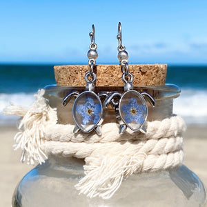 Forget Me Not Sea Turtle Earrings displayed on a cork attached to a bottle against a blurred beach background, perfect for beach-themed accessories.