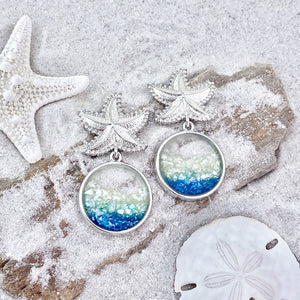 Glittering Ocean Starfish Earrings are displayed by being placed on top of a sand covered driftwood.