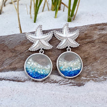 Load image into Gallery viewer, Glittering Ocean Starfish Earrings are displayed by being placed on top of a driftwood on the sand.