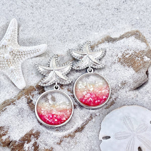 Glittering Ocean Starfish Earrings are displayed by being placed on top of a sand covered driftwood.