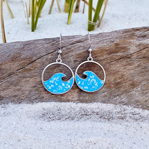 Glittering Wave Earrings are displayed by being placed on top of a driftwood on the sand.