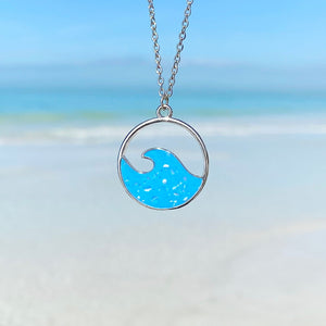 Glittering Wave Necklace is hanging up close with a beach background blurred.