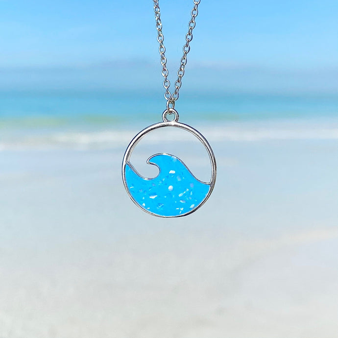 Glittering Wave Necklace is hanging up close with a beach background blurred.
