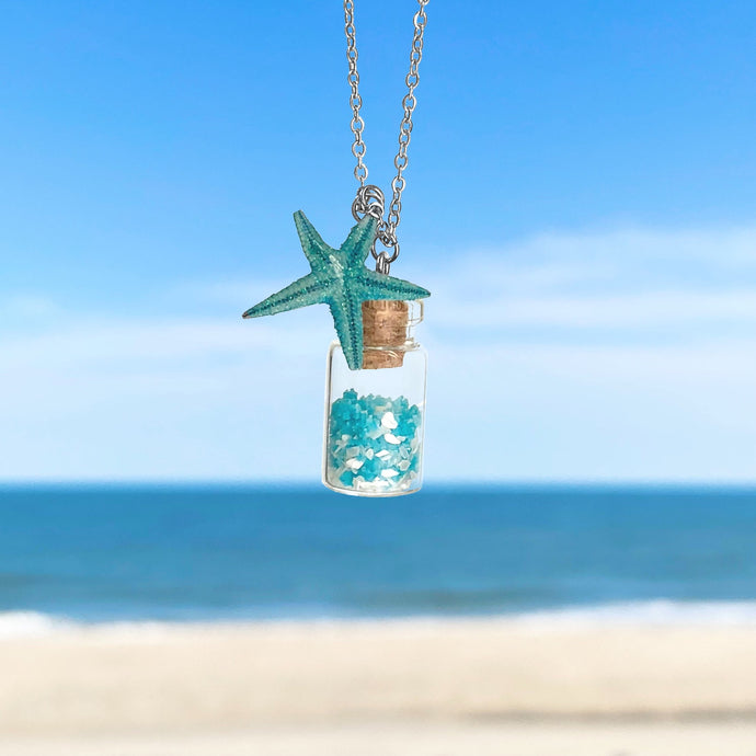 Glowing Sand Jar Necklace hanging close for a shot with a blurred beach background.