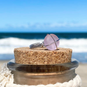 Hand-Wired Pink Sea Glass Ring is placed on top of a cork bottle against a blurred beach background.
