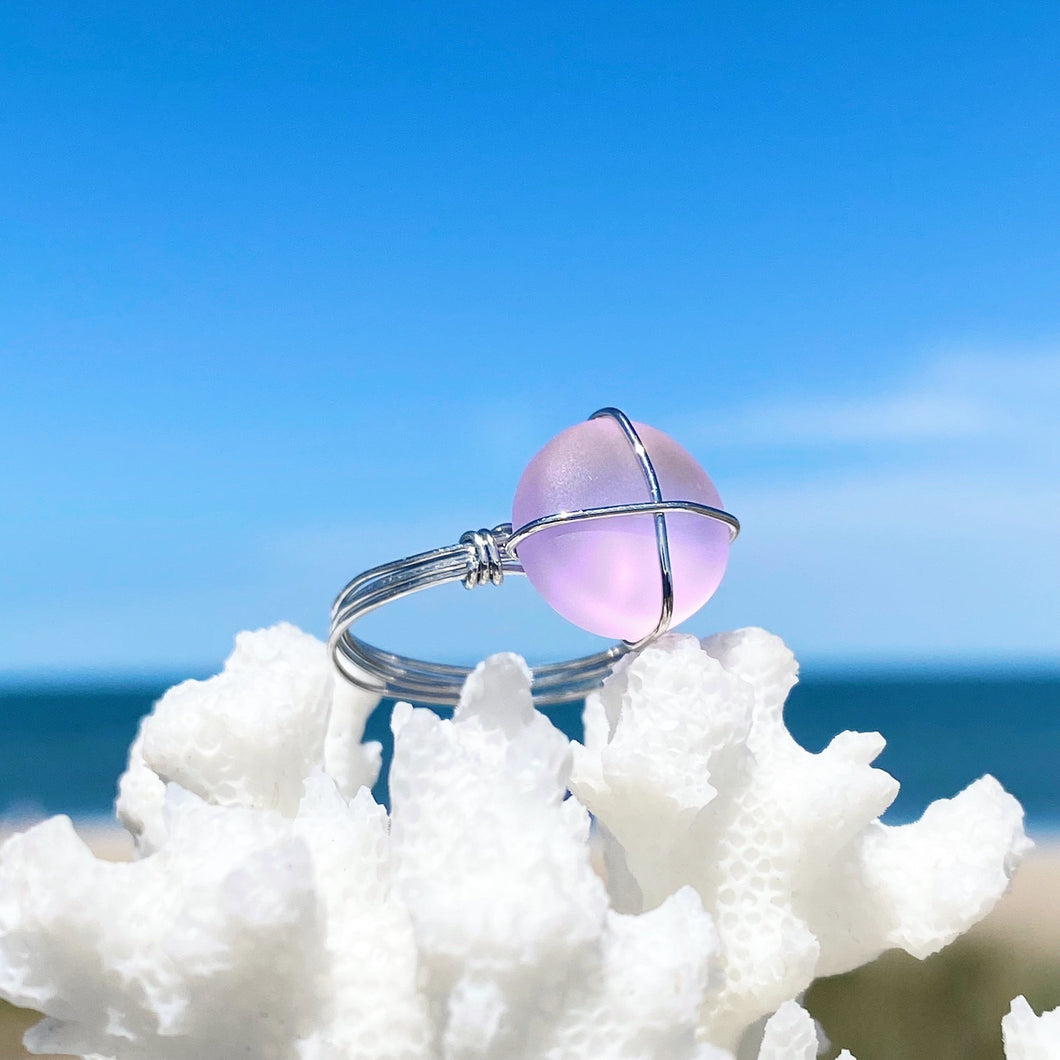 Hand-Wired Pink Sea Glass Ring is sitting on top of a dried white artificial coral against a blurred beach background.