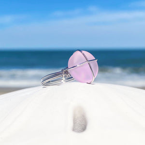 Hand-Wired Pink Sea Glass Ring is sitting on top of a white dome against a blurred beach background.