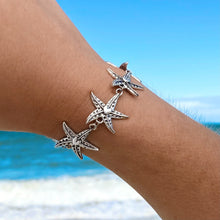 Load image into Gallery viewer, Happy Starfish Heart Bracelet displayed by being worn around a woman&#39;s wrist against a blurred beach background..