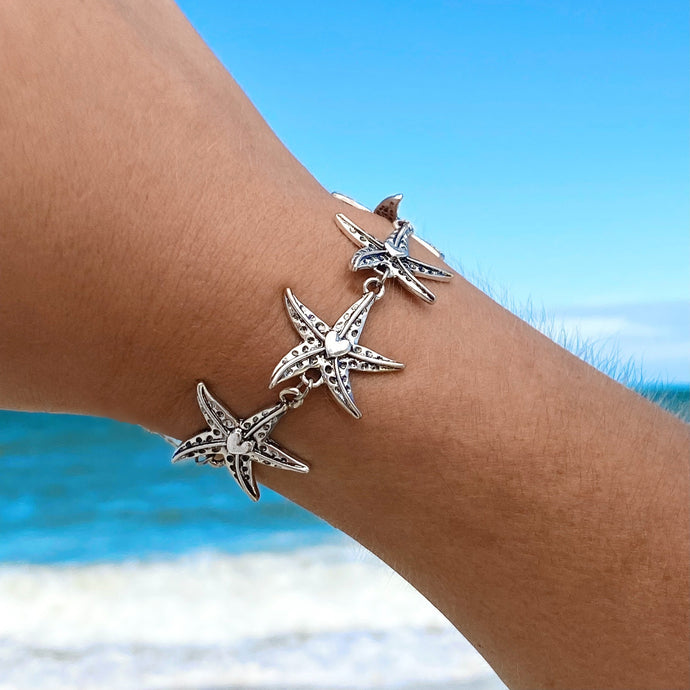 Happy Starfish Heart Bracelet displayed by being worn around a woman's wrist against a blurred beach background..