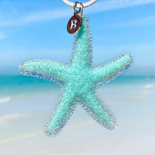 Load image into Gallery viewer, Happy Starfish Keychain hanging close for a shot with a blurred beach background.