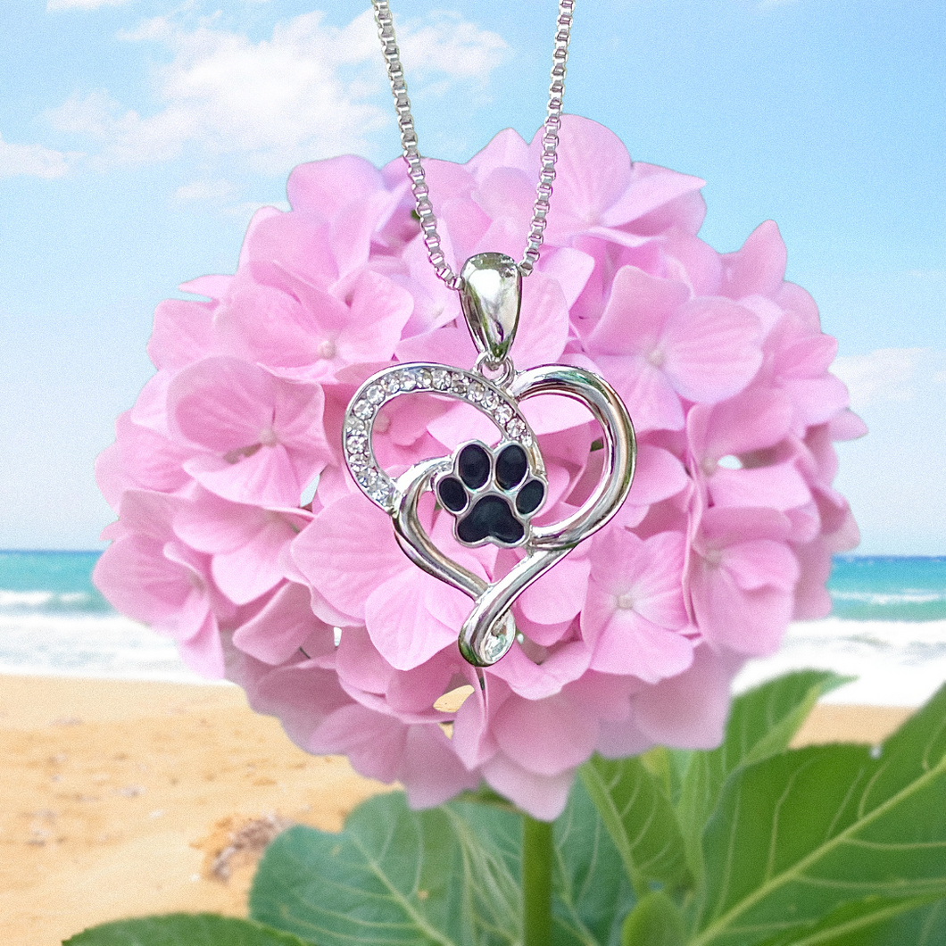 Heart Paw Necklace hanging close for a shot with a carnation pink flower in the background on the beach.