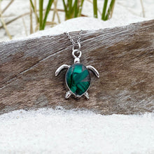 Load image into Gallery viewer, Malachite Sea Turtle Necklace is displayed by being placed on top of a driftwood on the sand.