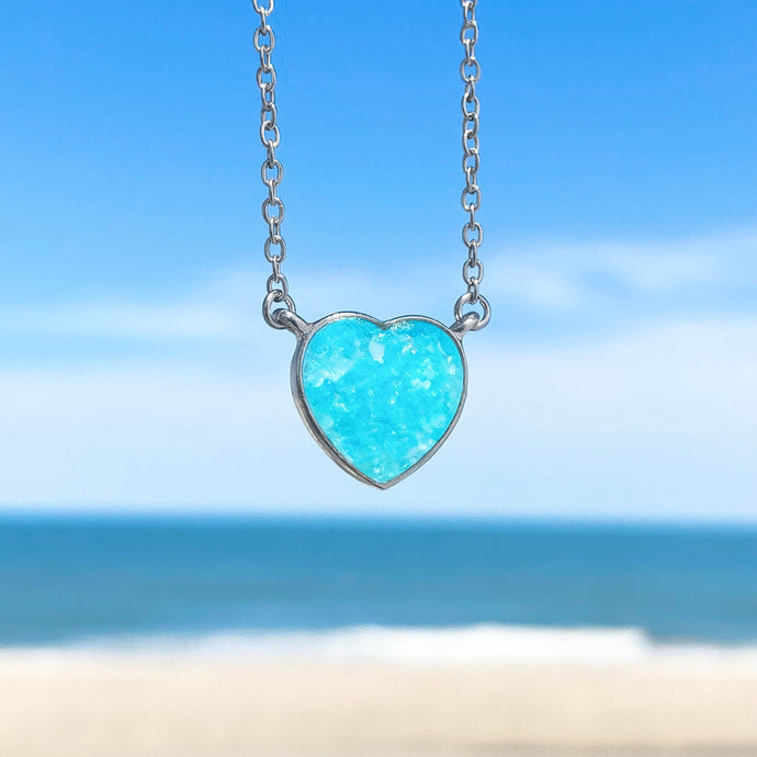 Ocean Treasure Sand Heart Necklace hanging in a close-up shot with a beach background blurred.