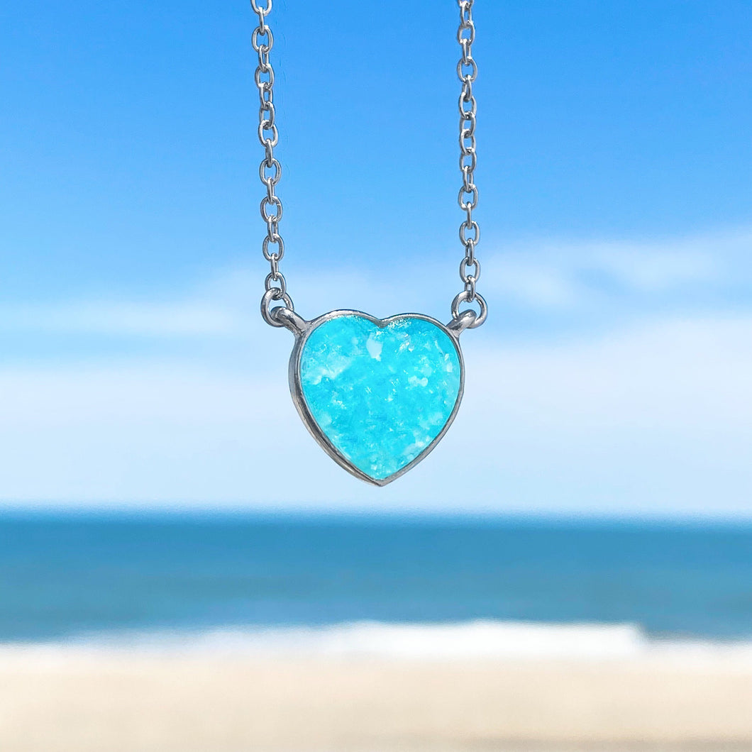 Ocean Treasure Sand Heart Necklace hanging in a close-up shot with a beach background blurred.
