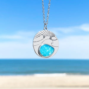 The Ocean Treasure Sand Wave Necklace is hanging up close with a blurry beach background.