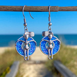 Opal Floatie Girl Earrings hanging on a stick for a shot with a blurred beach background.