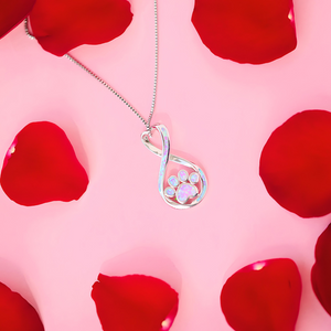 Opal Infinity Love Paw Necklace in Pink displayed on top of a pink surface with rose petals around it.