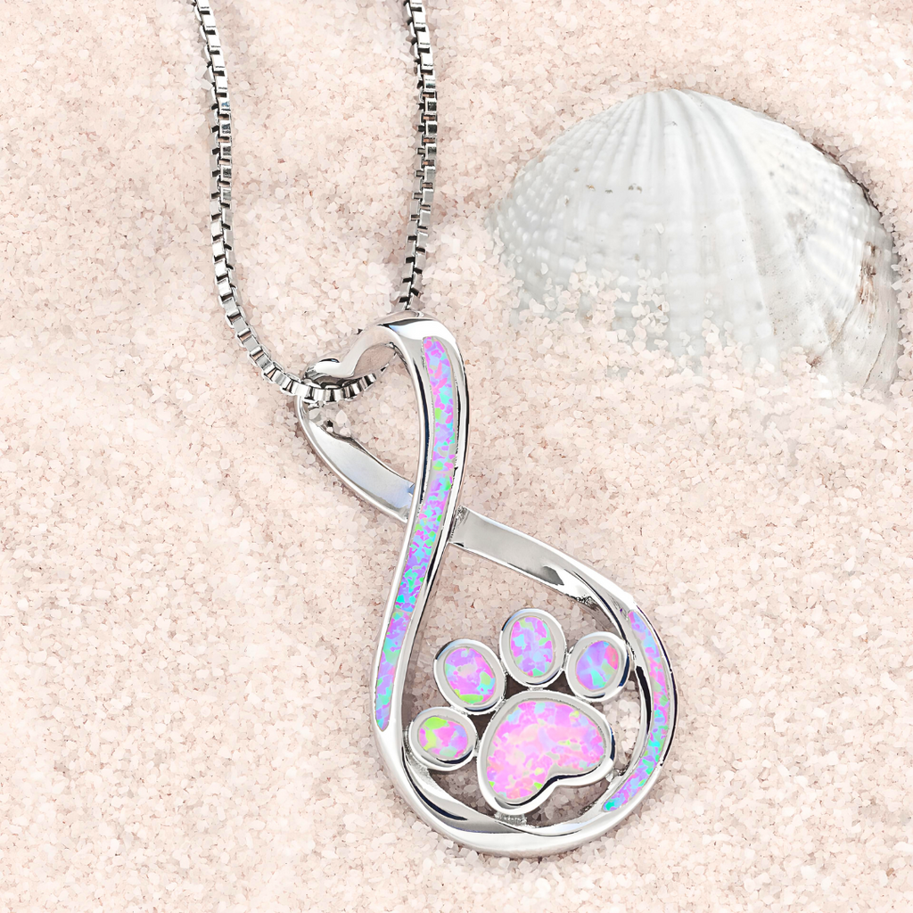 Opal Infinity Love Paw Necklace in Pink is displayed on a sandy surface.