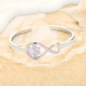 Opal Infinity Love Paw Cuff Bracelet in Pink, captured in a close-up shot on a beach shore.