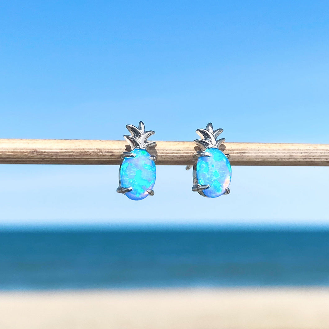 Opal Pineapple Stud Earrings are hanging on a stick in a close-up shot with a beach background blurred.