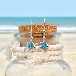 Opal Sea Turtle Flower Earrings displayed on a cork attached to a bottle against a blurred beach background, perfect for beach-themed accessories.
