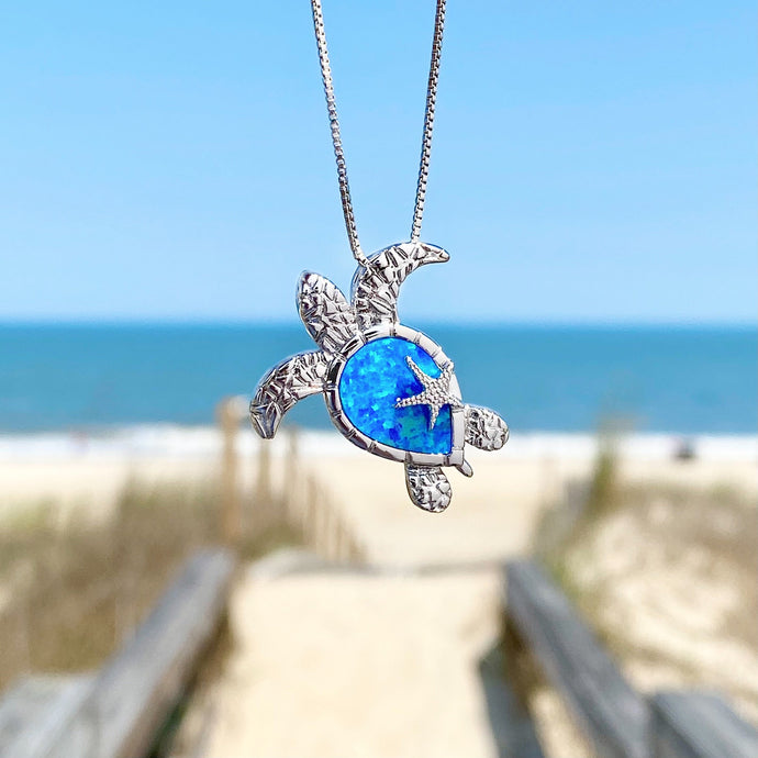 Opal Sea Turtle Starfish Necklace hanging close for a shot with a blurred beach background.