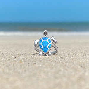 Opal Swimming Sea Turtle Ring is showcased on the sandy shore against a blurred beach background.