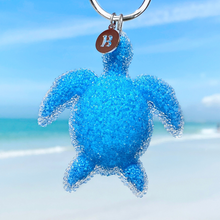 Load image into Gallery viewer, Playful Sea Turtle Keychain hanging close for a shot with a blurred beach background.