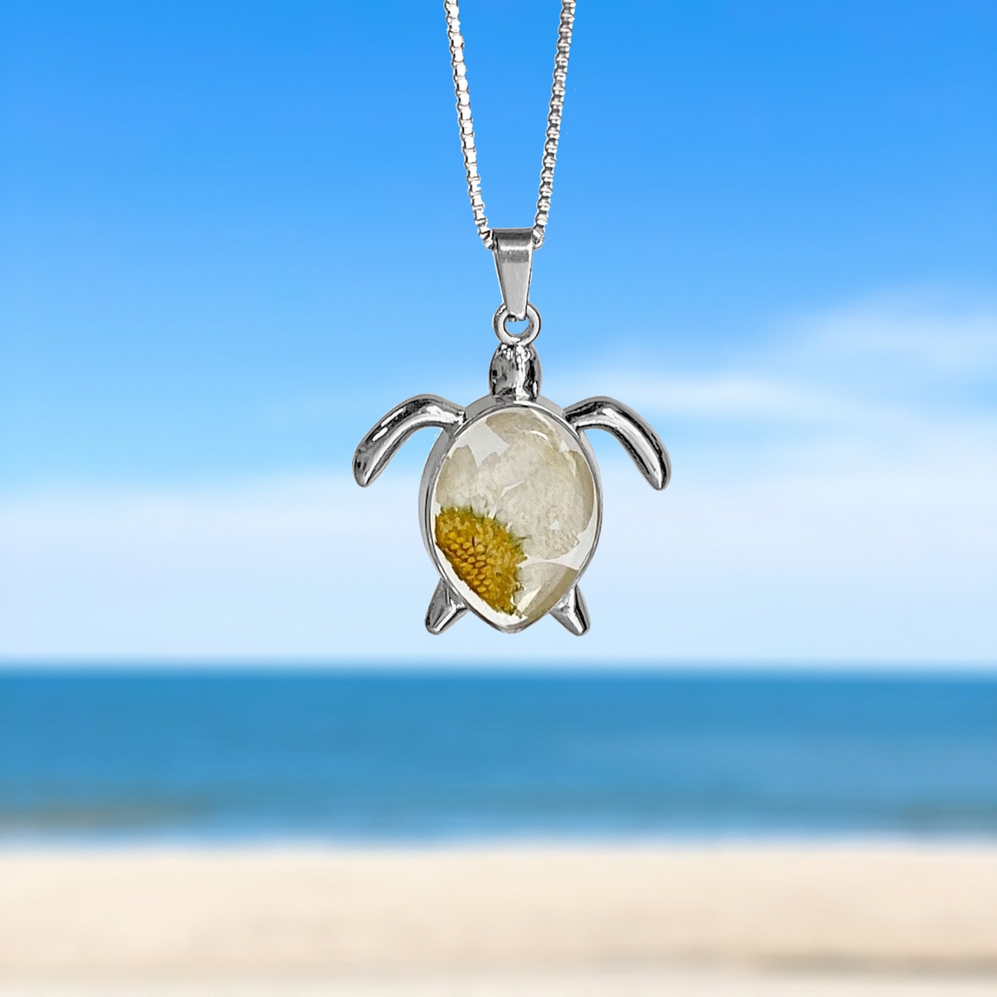 Pressed Daisy Sea Turtle Necklace hanging against a blurred beach background, perfect for beach-themed accessories.
