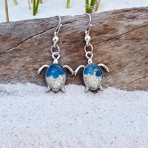 Sand Sea Turtle Earrings are displayed by being placed on top of a driftwood on the sand.