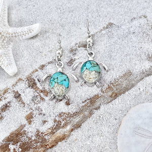 Sand Sea Turtle Earrings are displayed by being placed on top of a sand covered driftwood.
