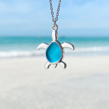 Load image into Gallery viewer, Sea Glass Sea Turtle Necklace in Sky Blue hanging close for a shot with a blurred beach background.