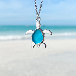 Sea Glass Sea Turtle Necklace in Sky Blue hanging close for a shot with a blurred beach background.