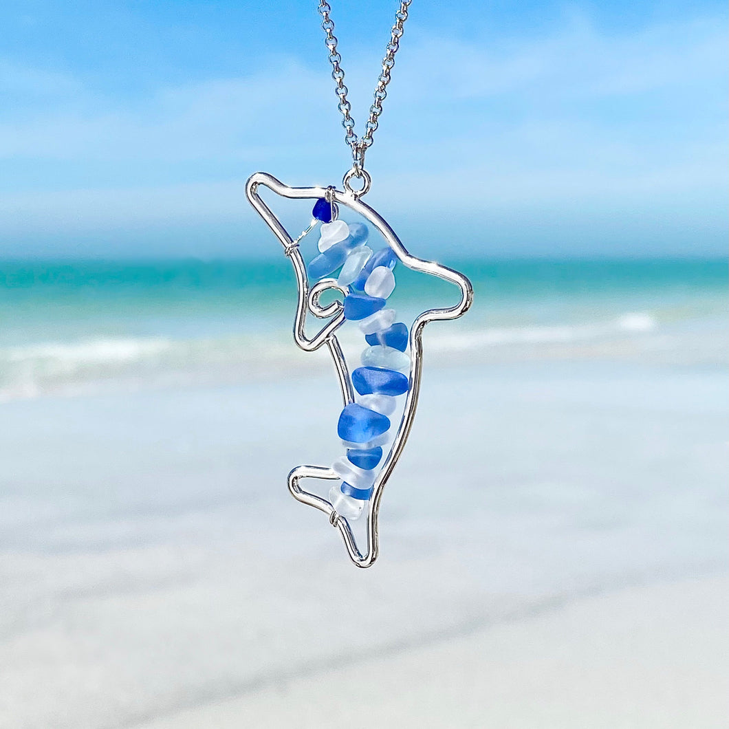 Stacked Sea Glass Dolphin Necklace hanging close for a shot with a blurred beach background.