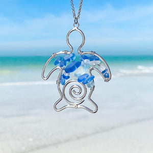 The Stacked Sea Glass Sea Turtle Necklace is hanging up close with a blurred beach background.