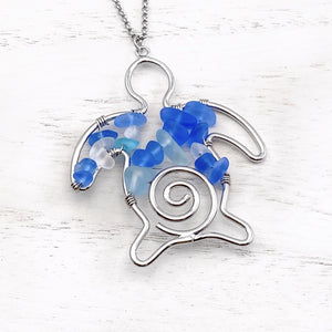 The Stacked Sea Glass Sea Turtle Necklace is displayed on a white wooden surface.
