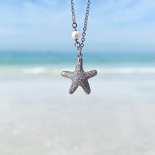 Load image into Gallery viewer, Starfish Hidden Pearl Necklace hanging close for a shot with a blurred beach background.
