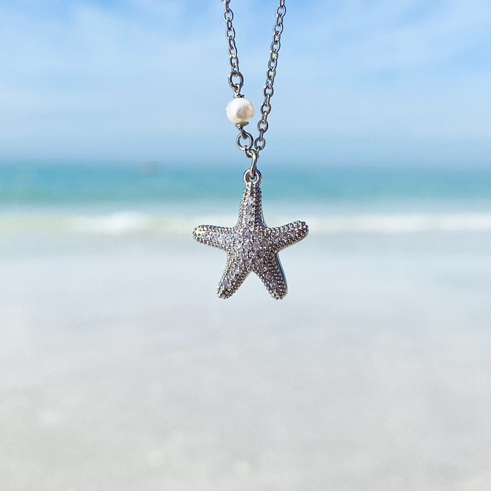 Starfish Hidden Pearl Necklace hanging close for a shot with a blurred beach background.