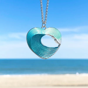 Wave Heart Necklace hanging in a close-up shot with a beach background blurred.