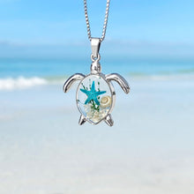 Load image into Gallery viewer, Deep in the Ocean Sea Turtle Necklace hanging close for a shot with a blurred beach background.