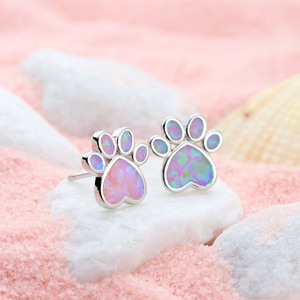 Opal Love Paw Studs in Pink resting on a white stone in a pink sandy environment.