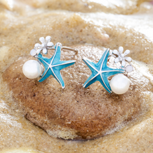 Load image into Gallery viewer, Starfish Pearl Studs are placed on top of a rock which is slightly buried in the sand.