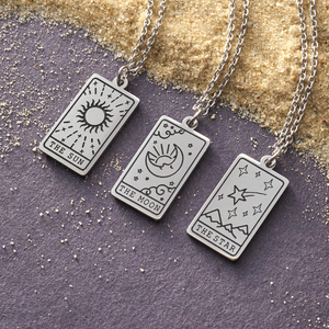 Necklaces featuring Moon Tarot, Sun Tarot, and Star Tarot Cards displayed on a gray surface with sandy grains.